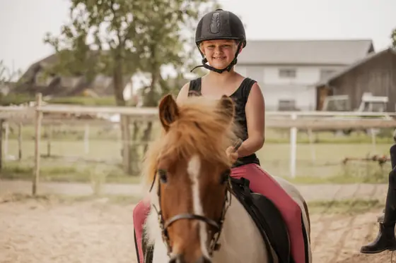 Ein Mädchen sitzt lachend auf einem Pony auf dem Reitplatz.